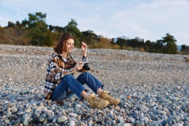 Kayalık bir plajda oturan sıradan bir kadın, açık hava manzarası, kot pantolon, ekose gömlek ve botlar, sakin bir manzara ve temiz deniz havası..