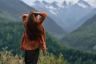 Kahverengi ceketli bir kadın engebeli dağlara ve orman yamaçlarına tepeden bakıyor. Gündüz boyunca görkemli dağlık arazide keşif ve sessiz bir yansıma yaratıyor.