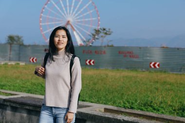 Dönme dolabı olan bir parkta kazak ve kot giyen bir kadının günlük duruşu, açık hava eğlence sahnesi, güneşli bir gün, şehir oyun havası, taze ve ulaşılabilir bir hava, neşeli bir ortam.