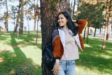 Ağaçlara yaslanan, parkta doğanın tadını çıkaran, kot pantolon ve örgü bluzlu, öğleden sonra ışığı ve gölgesi olan, sakin manzaralı, sağlıklı yaşam tarzı olan bir kadın.