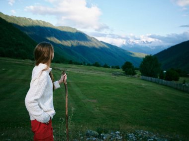 Samimi bir süveter giyen yalnız bir yürüyüşçüyle açık bir kırsal bölge manzarası. Yuvarlanan tepelere, çayırlara ve mavi gökyüzüne bakıyor. Macera, doğa ve özgürlükleri aktarıyor.