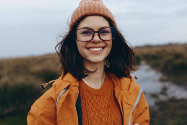 Gözlüklü ve kahverengi şapkalı mutlu kadın dışarıda sonbaharın tadını çıkarıyor. Turuncu ceket ve kazak giymiş, doğal çevrede gülümsüyor, yaşam tarzı ve mevsimsel moda kavramı.