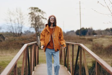 Mutlu kadın yaşam tarzı, ahşap köprüde, gözlüklü ve bereli gündelik kıyafetlerle duruyor. Doğal arka plan, rahat hava ve ılık sonbahar renklerine sahip açık hava sahnesi..