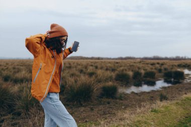 Genç kadın yaşam tarzı, günlük kıyafetlerle dışarıda, sulak çayırların yanında yürüyor, akıllı telefon tutuyor ve kulaklıkla müzik dinliyor, rahat ve aktif ruh halini gösteriyor..