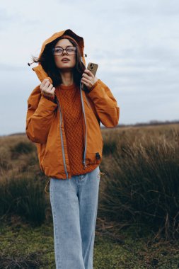 Açık hava yaşam tarzı, turuncu ceketli ve gözlüklü genç bir kadının cep telefonuyla sahada durması. Gündelik sonbahar modası, doğal arka plan, rahat ruh hali ve modern teknoloji konsepti..