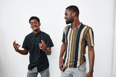 Esmer tenli iki Afro-Amerikan adam günlük kıyafetlerle poz veriyor ve gülümsüyor. Biri siyah gömlekli, diğeri çizgili kısa kollu gömlekli, ikisi de beyaz arka planda duruyor..