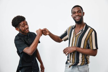 Siyah tenli, sıradan gömlekli Afro-Amerikalı erkekler yumruk tokuşturup gülüyorlar. Beyaz stüdyo arka planında iki genç siyah erkek arasındaki arkadaşlık, neşe ve sosyal etkileşim.