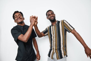 Siyah tenli, sıradan gömlekli, elini sıkan ve gülümseyen Afrikalı etnik erkekler. Genç siyahi erkekler beyaz stüdyo geçmişinde dostluk, neşe ve pozitif bir bağ gösteriyorlar..