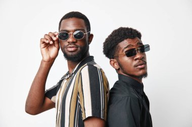 Siyah tenli, güneş gözlüklü ve arka arkaya giyinmiş iki Afrikalı adamın portresi. Moda tarzı ve beyaz stüdyo geçmişi konusunda kendine güveni olan genç siyahi erkekler..