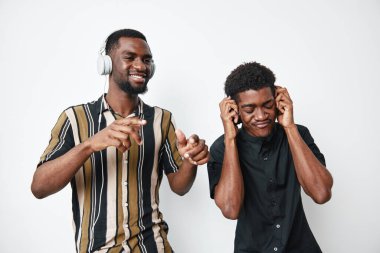 Koyu tenli Afro-Amerikalı erkekler kulaklıkla müzik dinliyor, günlük kıyafetlerle dans ediyor ve gülümsüyor, parlak stüdyo ortamında eğleniyor ve sesten keyif alıyorlar..