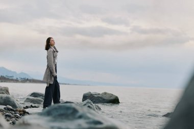 Uzun bir palto giymiş genç bir kadın uzak mesafedeki dağlarla sakin denize bakan kayalık sahilde duruyor. Yalnızlık, doğa ve barışçıl doğa manzarası kavramı..
