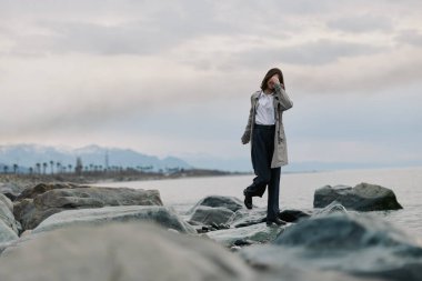Paltolu ve siyah pantolonlu bir kadın sakin deniz kıyısında ıslak kayalarda dikkatle yürür. Bulutlu gökyüzü ve uzak dağlar kıyı manzarasına barışçıl ve yansıtıcı bir hava katar..