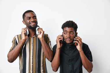 Koyu tenli, telefon ve kulaklıklı, sıradan kıyafetlerle poz veren Afrikalı genç adamlar. Beyaz stüdyo geçmişi olan teknoloji, bağlantı ve yaşam tarzı konsepti.