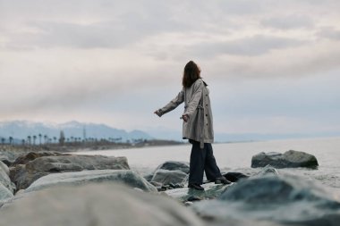 Uzun bir palto giyen bir kadın deniz kenarındaki kayaların üzerinde duruyor kolları açık, bulutlu gökyüzüne bakıyor. Huzurlu okyanus ve uzak dağlar sakin bir doğal manzara yaratır..