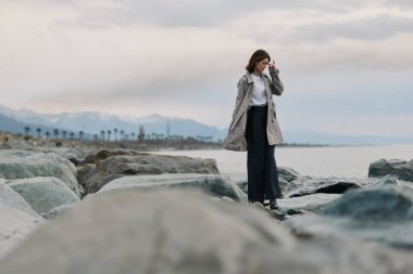 Okyanus kıyısındaki kayalık sahillerde palto giymiş, uzaklardaki dağlar ve palmiye ağaçları olan bir kadın. Bulutlu gökyüzü karamsar bir atmosfer yaratır. Barışçıl açık hava yaşam tarzı kavramı.