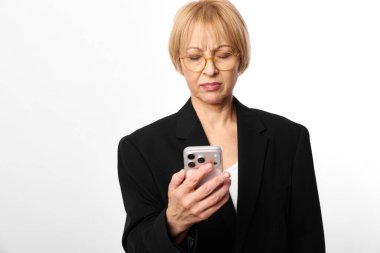 Gözlüklü ve siyah ceketli olgun bir kadın beyaz arka plandaki akıllı telefon ekranına bakıyor. Teknoloji kullanımı, iletişim, iş ve dijital bağlantı kavramı.