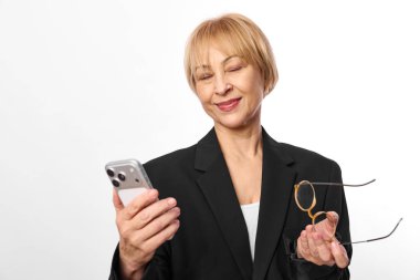 Siyah ceketli, kısa sarı saçlı, kendinden emin orta yaşlı kadın akıllı telefon ve gözlük tutuyor, gözleri kapalı gülümsüyor. Dijital iletişim, yaşam tarzı ve pozitif ruh hali kavramı.