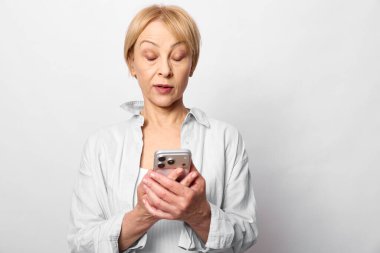 Beyaz bluz giyen yetişkin bir kadın telefonu iki eliyle tutup ekrana bakıyor. Tarafsız bir geçmişi olan bir stüdyo fotoğrafı. Mobil teknoloji ve reaksiyon kavramı.