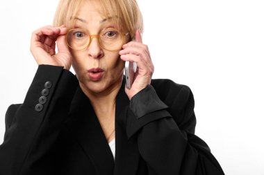 Gözlüklü ve siyah ceketli orta yaşlı bir kadın telefonu kulağına tutuyor, şaşırmış bir ifadeyle gözlük çerçevesine dokunuyor. Beyaz arka plan. İletişim, şok ve tepki kavramı.