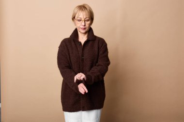 Koyu kahverengi ceketli ve beyaz pantolonlu orta yaşlı bir kadın bej arka planda elleri birarada duruyor. Sıradan moda, tarz, sonbahar kıyafetleri ve portre kavramı..