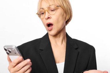 Kısa saçlı, siyah ceketli, şok olmuş orta yaşlı bir kadın akıllı telefona bakar ve beyaz arka planda elini kaldırır. Sürpriz ve duygu kavramı.