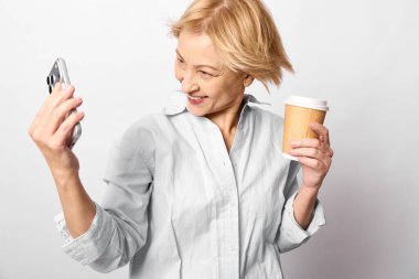 Beyaz gömlek giyen mutlu sarışın kadın kahveyi seviyor ve beyaz arka planda akıllı telefonlu selfie çekiyor. Günlük yaşam tarzı kavramı, sabah rutini, teknoloji ve pozitif ruh hali..