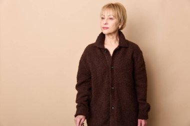 Kısa sarı saçlı düşünceli olgun bir kadın bej arka planda duran kahverengi yün bir ceket giyer. Yaşlanma konsepti, gündelik moda, yaşam tarzı ve yaşlı yetişkinler için öz yansıma..