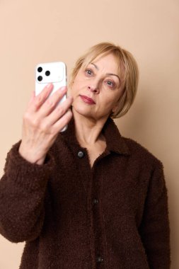Kısa sarı saçlı, kahverengi tüylü ceketli orta yaşlı kadın akıllı telefonlu selfie çekiyor. Bej arkaplan ve yumuşak ışıklandırma teknolojiyle gündelik, modern portre konsepti oluşturur.