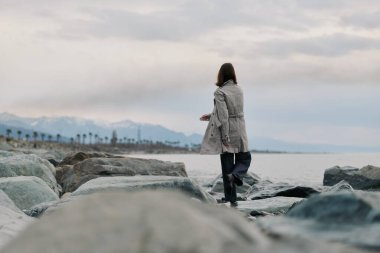 Sıcak bir palto giyen bir kişi uzak mesafeden görünen dağlar ve palmiye ağaçları ile deniz kıyısında büyük kayalar boyunca yürür. Yalnızlık, doğa, seyahat ve barışçıl açık hava keşfi kavramı..