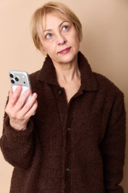 Sıcak kahverengi kazak giyen düşünceli orta yaşlı kadın akıllı telefonu tutuyor ve düşünceli bir ifadeyle yukarı bakıyor. Teknoloji ve yansıma odaklı bej arka planda izole edilmiş.