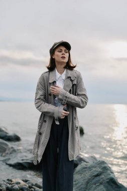 Gri paltolu ve şapkalı genç bir kadın gün batımında sakin deniz kenarında kayalık sahilde duruyor. Yumuşak doğal ışıklandırma, sakin bir ruh hali ve düşünceli bir ifadeyle sonbahar modasını vurgular..