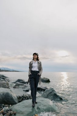 Gündelik sonbahar kıyafetleri içinde zarif bir kadın bulutlu gökyüzü olan kayalık sahilde poz veriyor. Moda, doğa, seyahat ve sakin atmosfer kavramı yumuşak doğal aydınlatma ve huzurlu ruh hali.