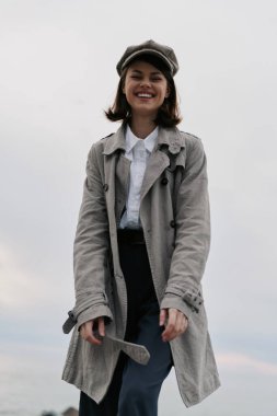 Açık havada gri tüvit ceket ve şapka giyen neşeli genç bir kadın. Sonbahar modası, gündelik tarz ve neşeli ifade bulutlu arka plan ve rahat duruş ile doğal ışık altında yakalanır..