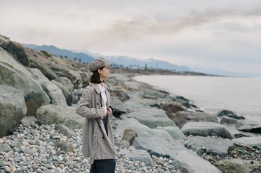 Gri paltolu ve şapkalı kadın okyanus kıyısındaki taşlı sahilde duruyor. Bulutlu hava ve dağ arka planı, barışçıl yalnızlığı ve doğa ile bağlantıyı vurguluyor..
