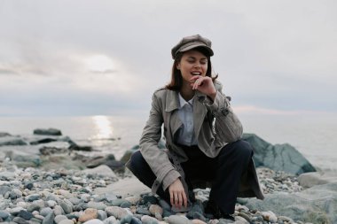 Kışlık palto ve haberci şapkası giyen gülümseyen genç kadın bulutlu gökyüzünün altında sakin suların yanında çakıl taşlı sahilde oturur, açık havada serin havanın tadını çıkarır..