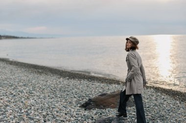 Gün batımında sakin denizin yanındaki kayalık sahilde gri bir ceket ve şapka giyen genç bir kadın. Yumuşak ışık ve bulutlu gökyüzü, dışarıdaki yalnızlığı, doğayı ve rahatlamayı vurgular..