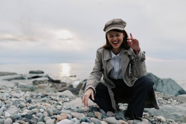 Şık bir palto ve gazeteci çocuk şapkası giyen mutlu genç bir kadın çakıl taşlı sahilde sakin suların yanında çömelmiş duruyor. Bulutlu gökyüzü ve doğal manzara açık havada rahat ve samimi bir atmosfer yaratır..