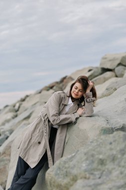 Örgülü kazak giyen sakin genç bir kadın sahilin yakınındaki büyük kayalara eğiliyor ve gözleri kapalı. Bulutlu gökyüzü ve doğal taşlar barışçıl bir atmosfer yaratır. Rahatlama, doğa ve konfor kavramı
