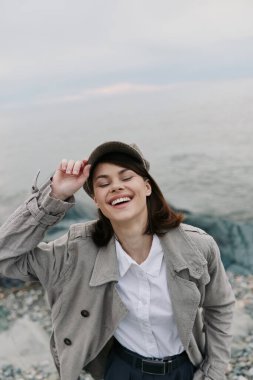 Kayalık sahilde ceketi ve tişörtü olan, gözleri kapalı gülümseyen bir kadın. Açık hava portresi rahat, kaygısız bir ifade ve doğal bir kıyı arkaplanı yakalar..