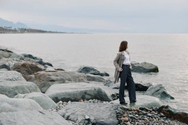 Kayalık sahilde sıradan bir kadın sakin suya ve bulutlu gökyüzüne bakar. Açık hava yalnızlığı, doğa keşfi ve doğal ışıklandırma ve sessiz atmosferli huzurlu kıyı çevresi kavramı..