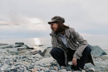 Paltolu ve şapkalı genç bir kadın bulutlu gökyüzünün altında sakin deniz kıyısında çömeliyor. Gündelik moda, sonbahar tarzı, doğal ışık ve derin düşüncelere dalma sahnesi..