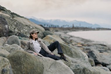 Şapka ve ceket giyen sıradan bir kadın deniz kenarındaki kayalık kıyı şeridinde görünür dağlarla oturuyor. Barışçıl açık hava rahatlama ve doğa keşfi kavramı.