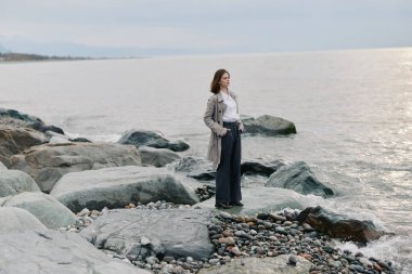 Paltolu ve koyu renk pantolonlu genç bir kadın kayalık sahilde deniz ufkuna bakıyor. Sakin su ve bulutlu gökyüzü sakin bir yansıma ve doğa kaçışı yaratıyor..