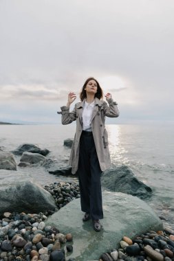 Uzun paltolu kendinden emin bir kadın bulutlu bir gökyüzünün altında sakin deniz suyunun yanındaki kayalık bir sahilde duruyor. Sakin atmosfer ve yumuşak ışık barışçıl ve yansıtıcı bir ruh hali yaratır..