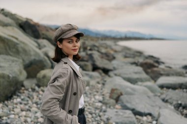 Kaliteli palto ve şapka giyen genç bir kadın kayalık sahilde deniz ve bulutlu gökyüzü ile poz veriyor. Doğal ışık, sonbahar moda tarzını ve plajda açık hava yaşam tarzını ön plana çıkarıyor..