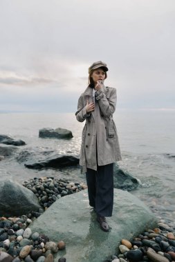Şık desenli bir palto giyen ve şapkalı bir kadın bulutlu bir gökyüzünün altında, etrafında taşlar ve çakıl taşları olan sakin bir denizin kenarındaki büyük taşların üzerinde duruyor. Moda ve doğa kavramı.