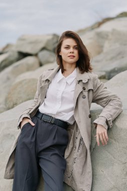 Kısa saçlı, desenli ceketli ve siyah pantolonlu genç bir kadın kayaya yaslanıyor. Tarafsız tonlarda ve rahat duruşlu açık hava moda fotoğrafı. Tesadüfi zarafet ve güven kavramı.