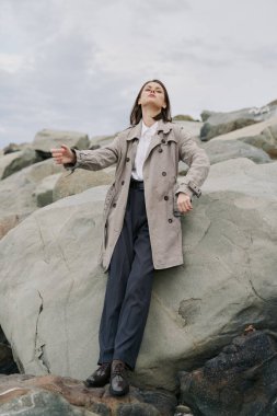 Uzun bir palto ve koyu renk pantolon giyen genç bir kadın, kolları hafifçe havada kayaların üzerinde duruyor. Bulutlu gökyüzü ve doğal taşlar huzur ve bağımsızlık havası yaratır..