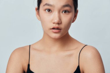 Pürüzsüz cilt ve az makyajlı koyu renk saçlı genç bir manken. Beyaz arka planda güzellik veya moda konsepti için ciddi bir yüz ifadesi olan ince kayışlı siyah bir bluz..