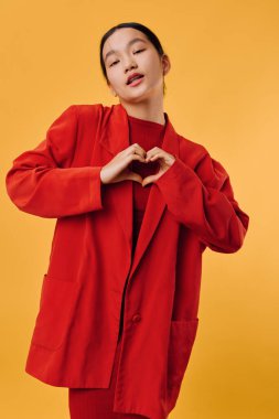 Elleriyle kalp sembolü yapan kırmızı ceketli, gülümseyen genç bir kadının portresi. Sıcak sarı arka planlı bir stüdyo çekimi, günlük kıyafetler ve dostça bir ifade, şefkat ve nezaket göstergesi..